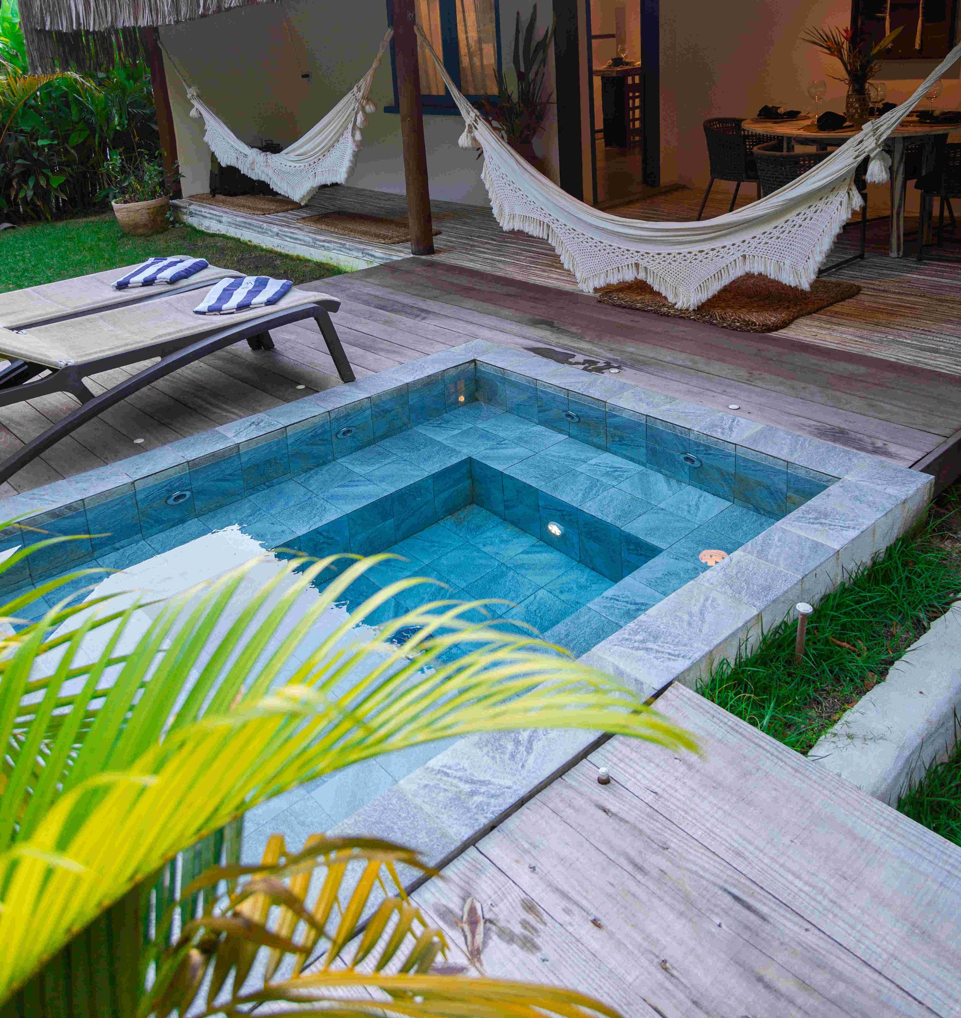Pátio tropical ensolarado com cadeiras de vime, rede e porta azul ao lado de um gramado verde.
