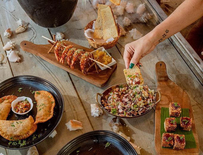 Vista aérea de uma mesa rústica com diversos aperitivos e bebidas, e uma mão servindo salada.