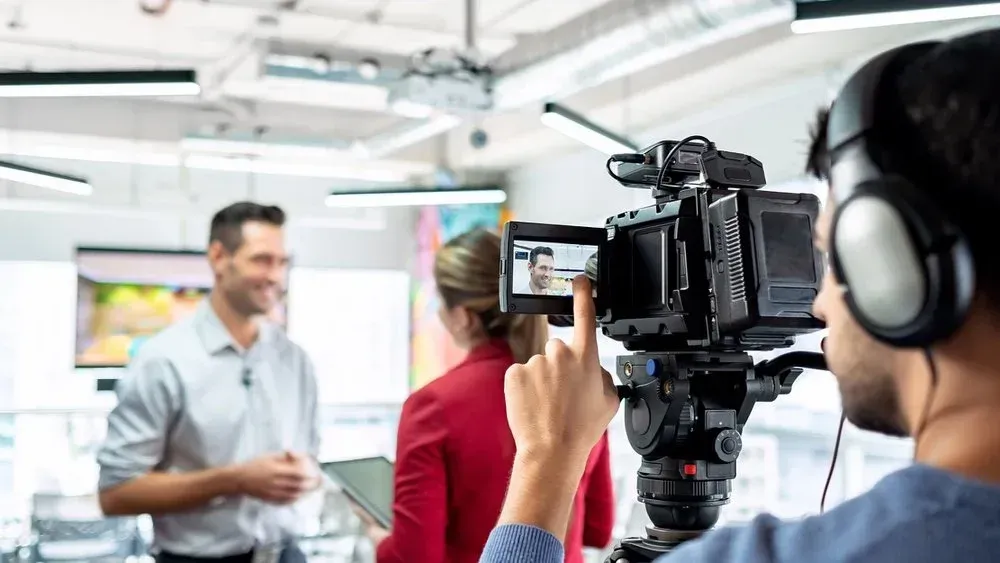 Videographer filming a man and woman in a bright studio. The camera operator is wearing headphones.