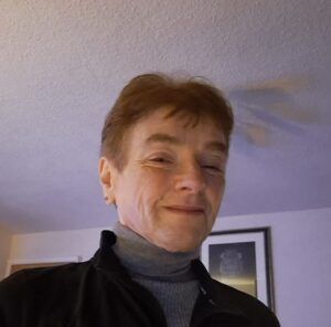 Smiling person indoors in a gray turtleneck and dark jacket, standing beneath a ceiling fan.