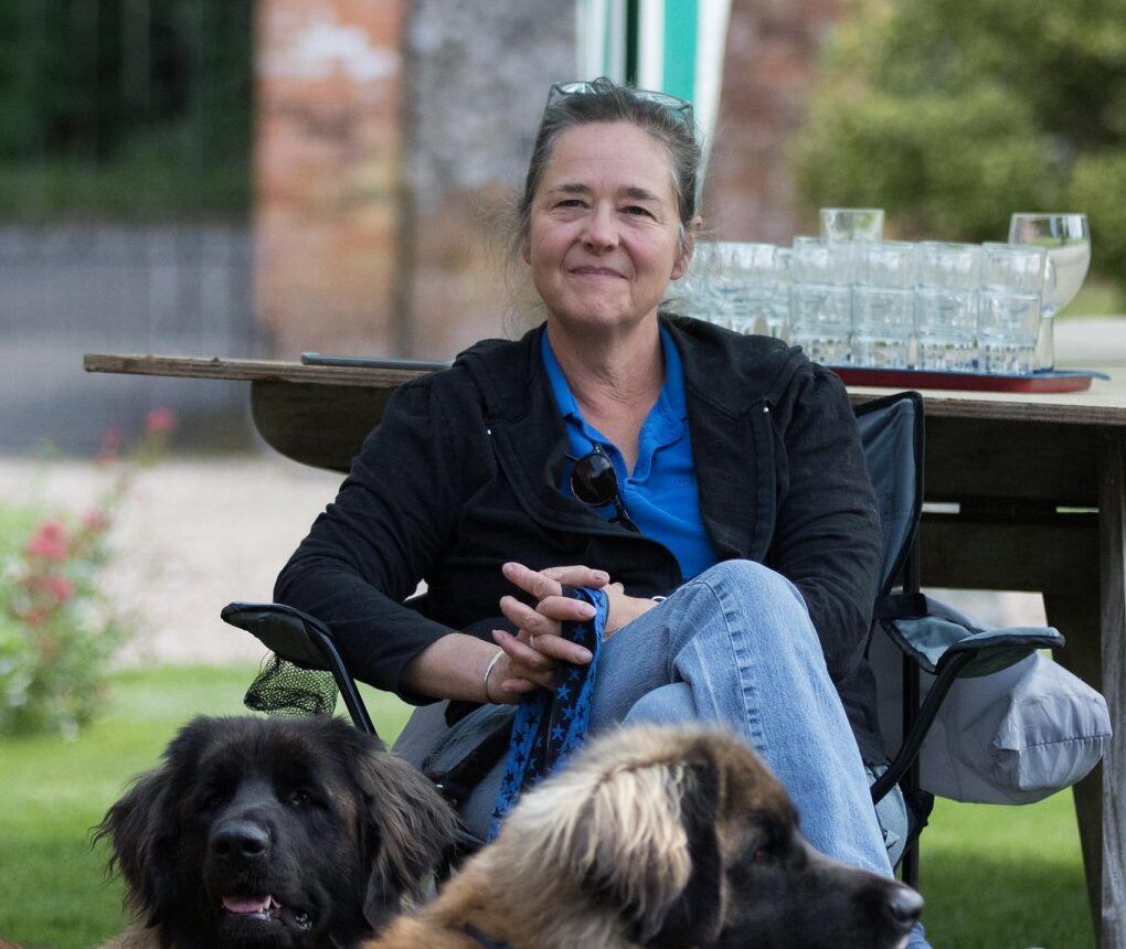 Woman sitting outdoors at a picnic table with two dogs in front, smiling at the camera