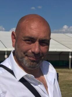 Close-up of a smiling bald man in a white shirt outdoors, with a white tent and blue sky behind him