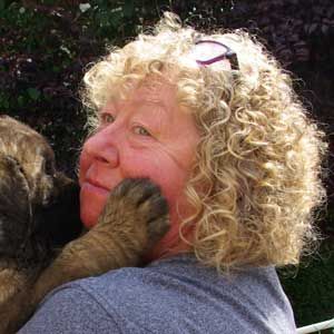 Woman with curly blonde hair being nuzzled by a dog outdoors