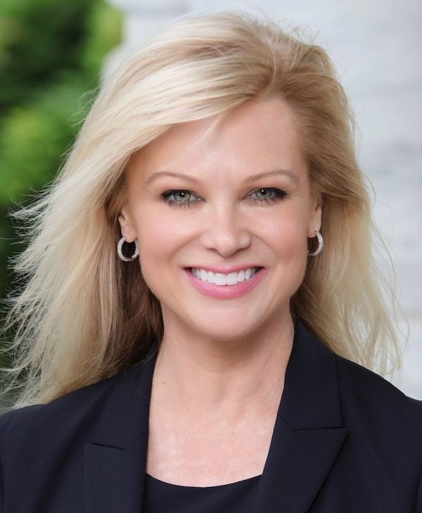 Lisa Wells in a black blazer with blonde hair and hoop earrings against a blurred outdoor background