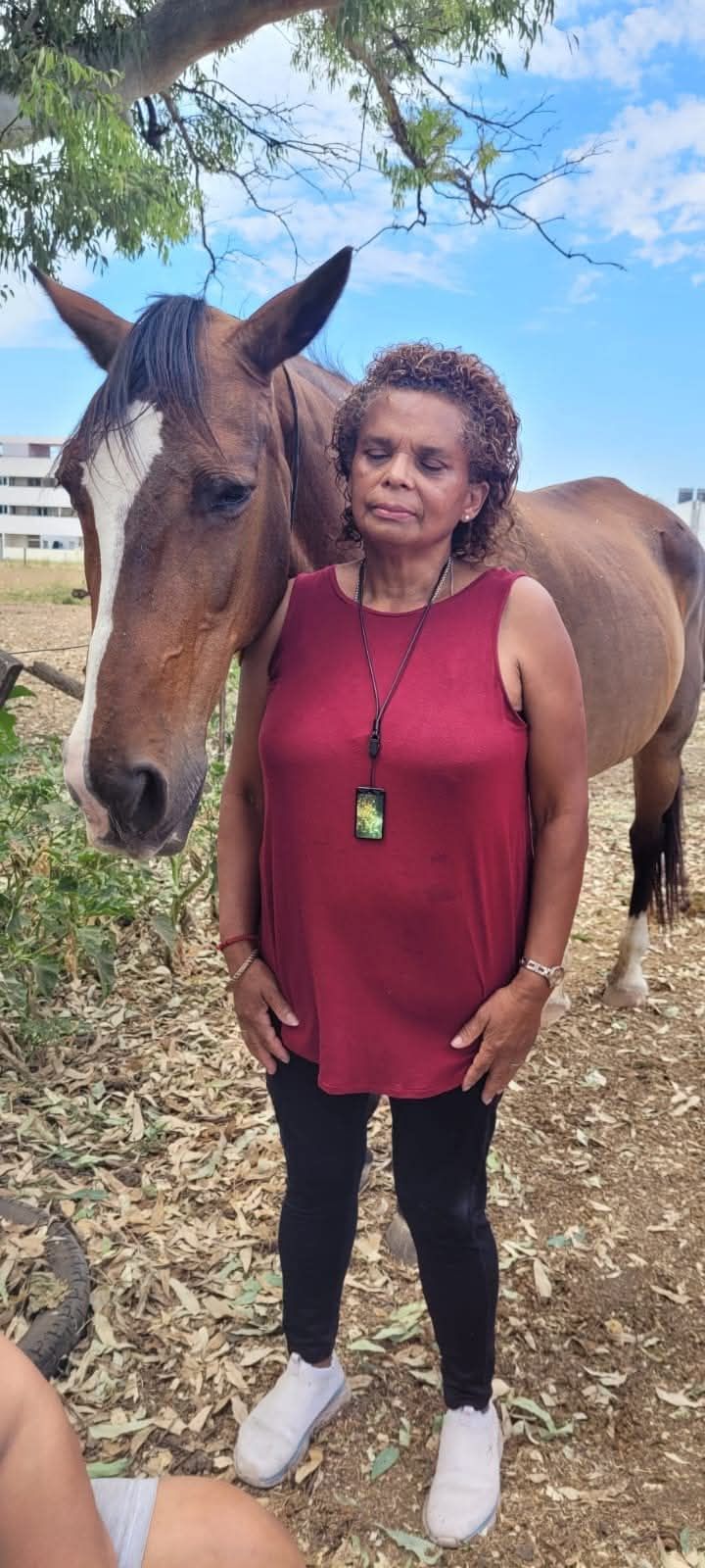 Mujer con blusa roja de pie junto a un caballo marrón al aire libre en un sendero boscoso.
