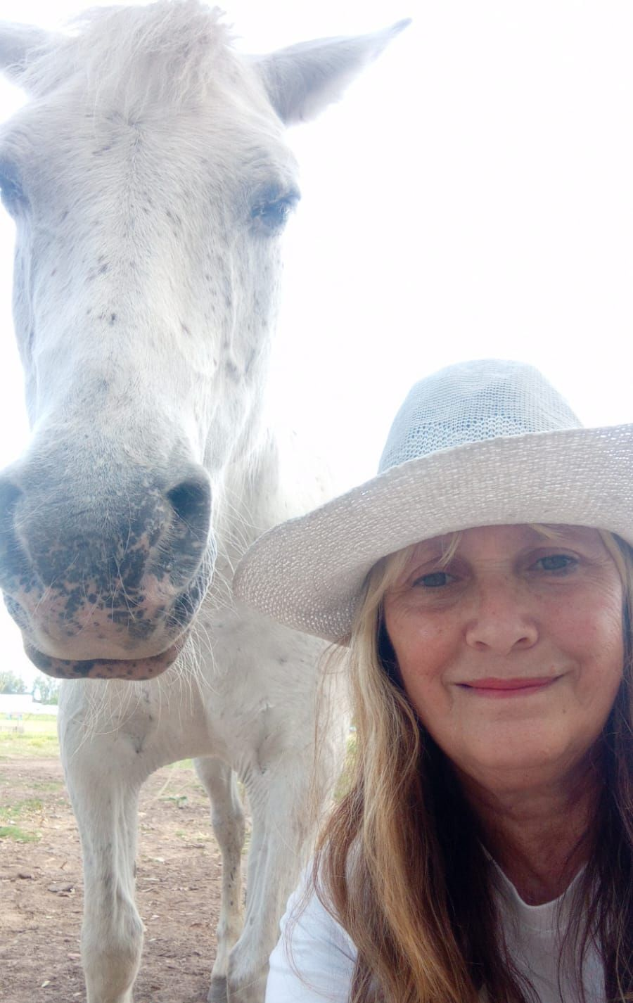 Mujer con sombrero blanco junto a un caballo blanco al aire libre, ambos mirando a la cámara.