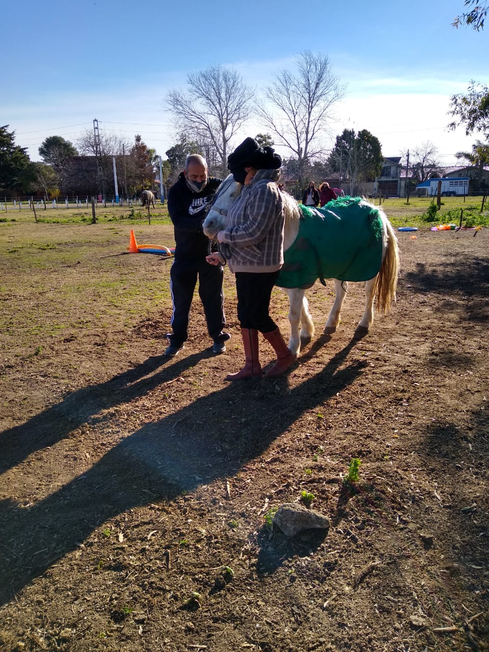 Dos personas ayudan a un pequeño caballo vestido con una manta verde en un campo de hierba en un día soleado.