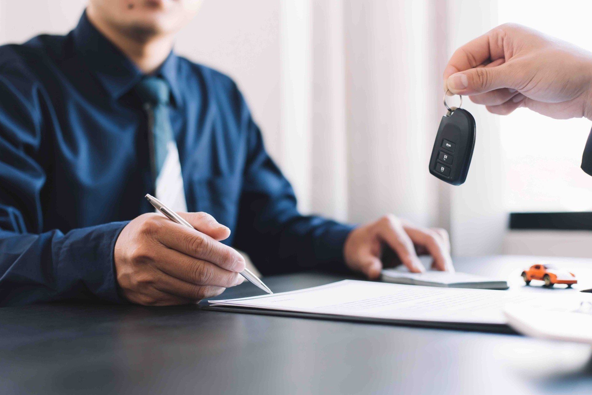 A man is sitting at a table signing a document while a man is holding a car key.