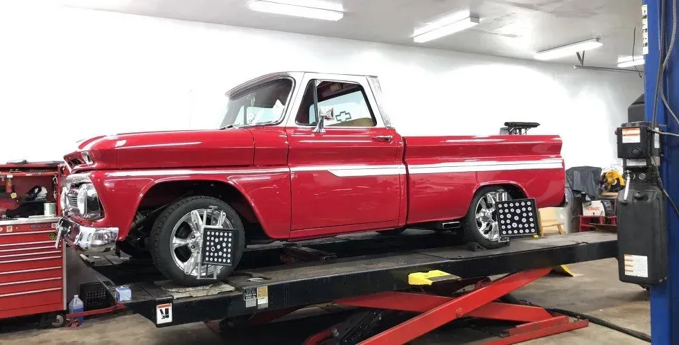 Red and white classic pickup truck on a lift, undergoing wheel alignment in a garage.