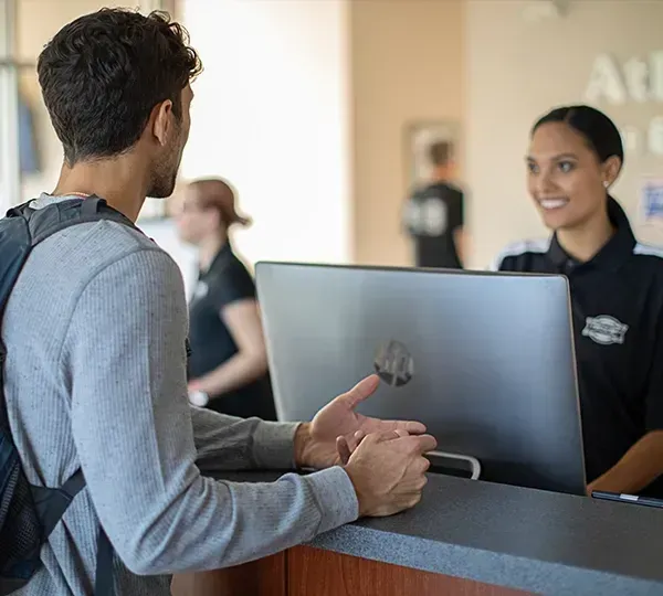 Man with backpack speaking to a woman at a desk with a computer.