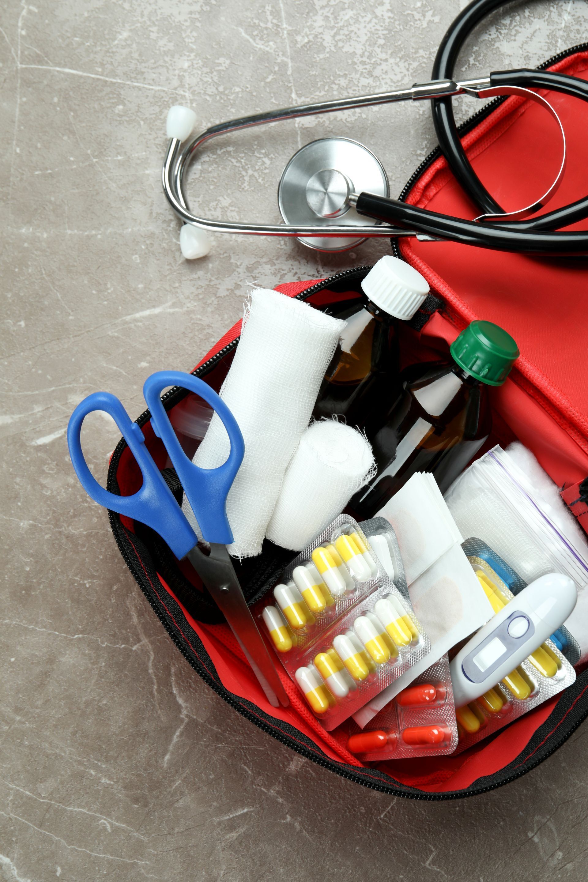 A first aid kit with a stethoscope , scissors , pills , bandages and a thermometer.
