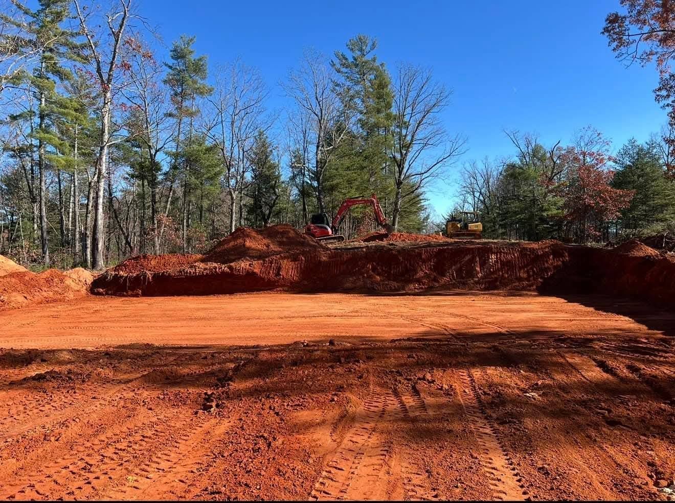 An excavated plot of red clay soil at a construction site with heavy machinery parked against a backdrop of trees.