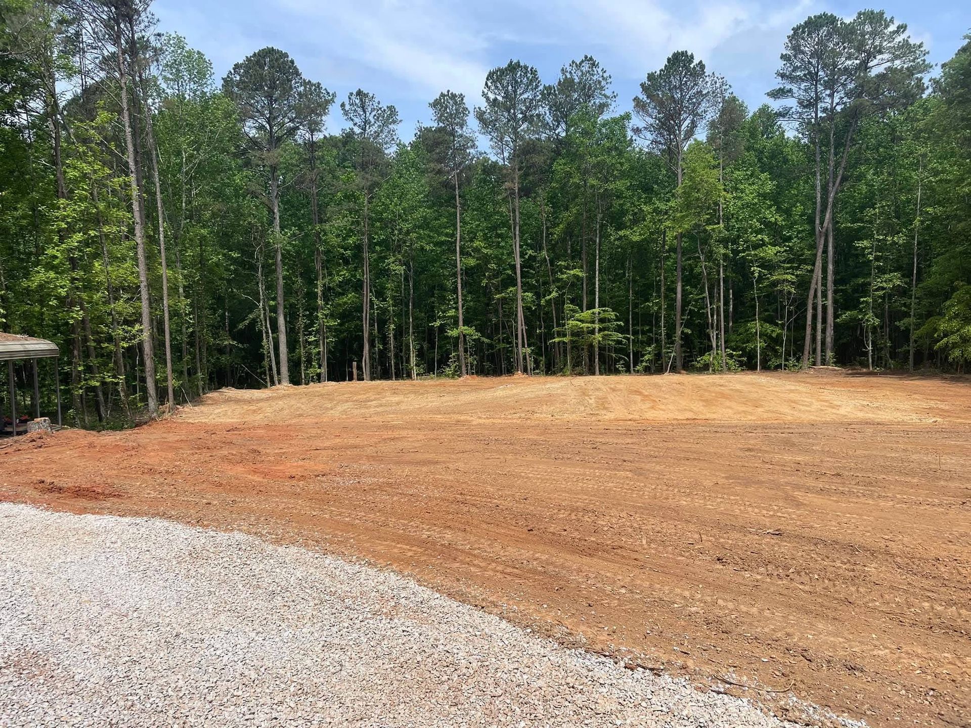 A patch of cleared, reddish dirt borders a gravel driveway, with a dense forest of tall green trees in the background.