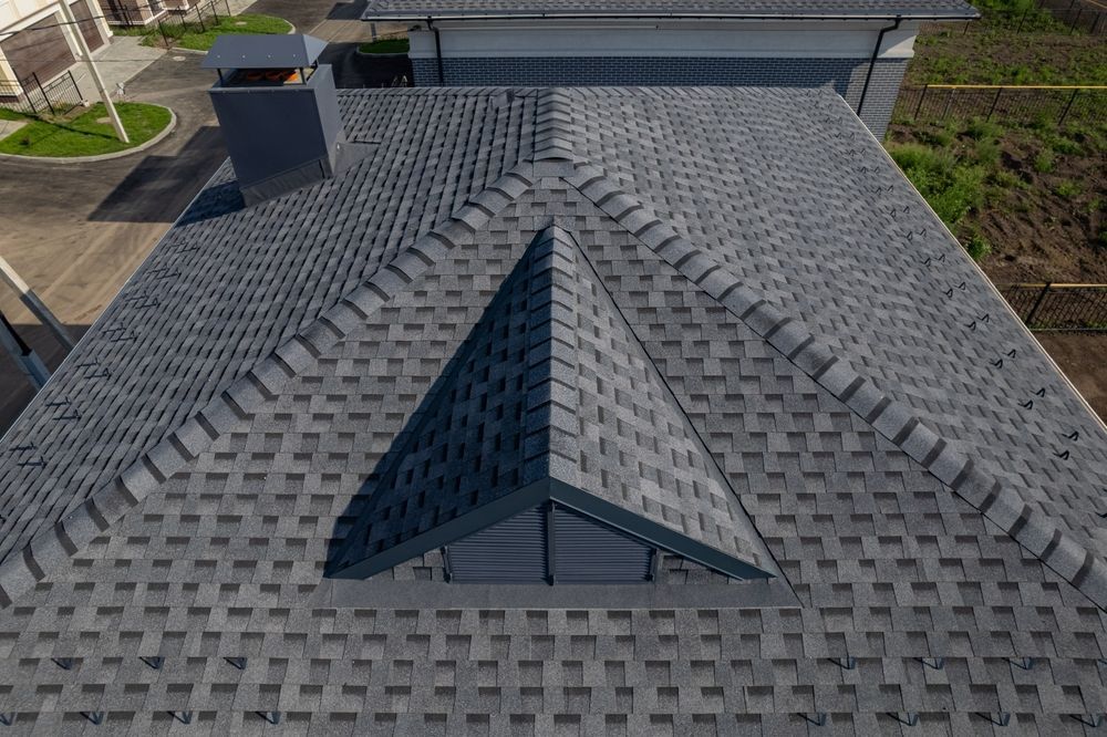 Overhead view of a house roof with dark gray asphalt shingles. A triangular dormer and chimney are visible.