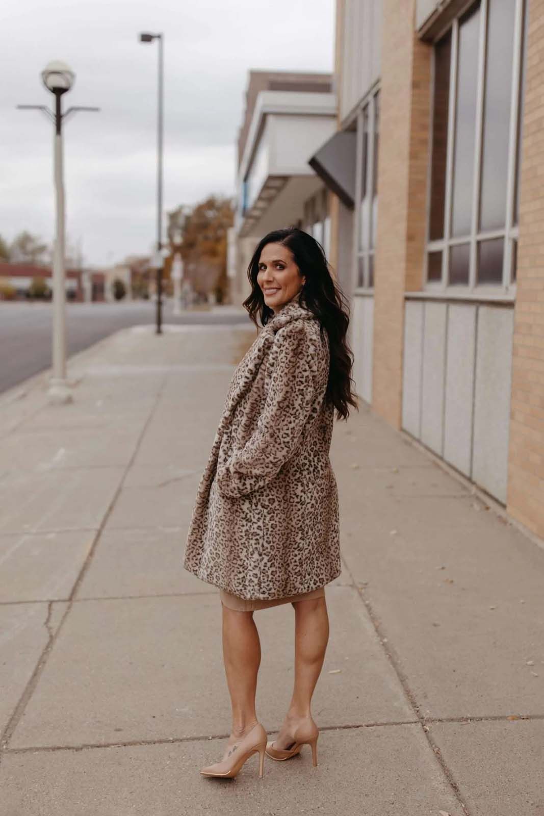 A woman is standing on a sidewalk wearing a leopard print coat and heels.