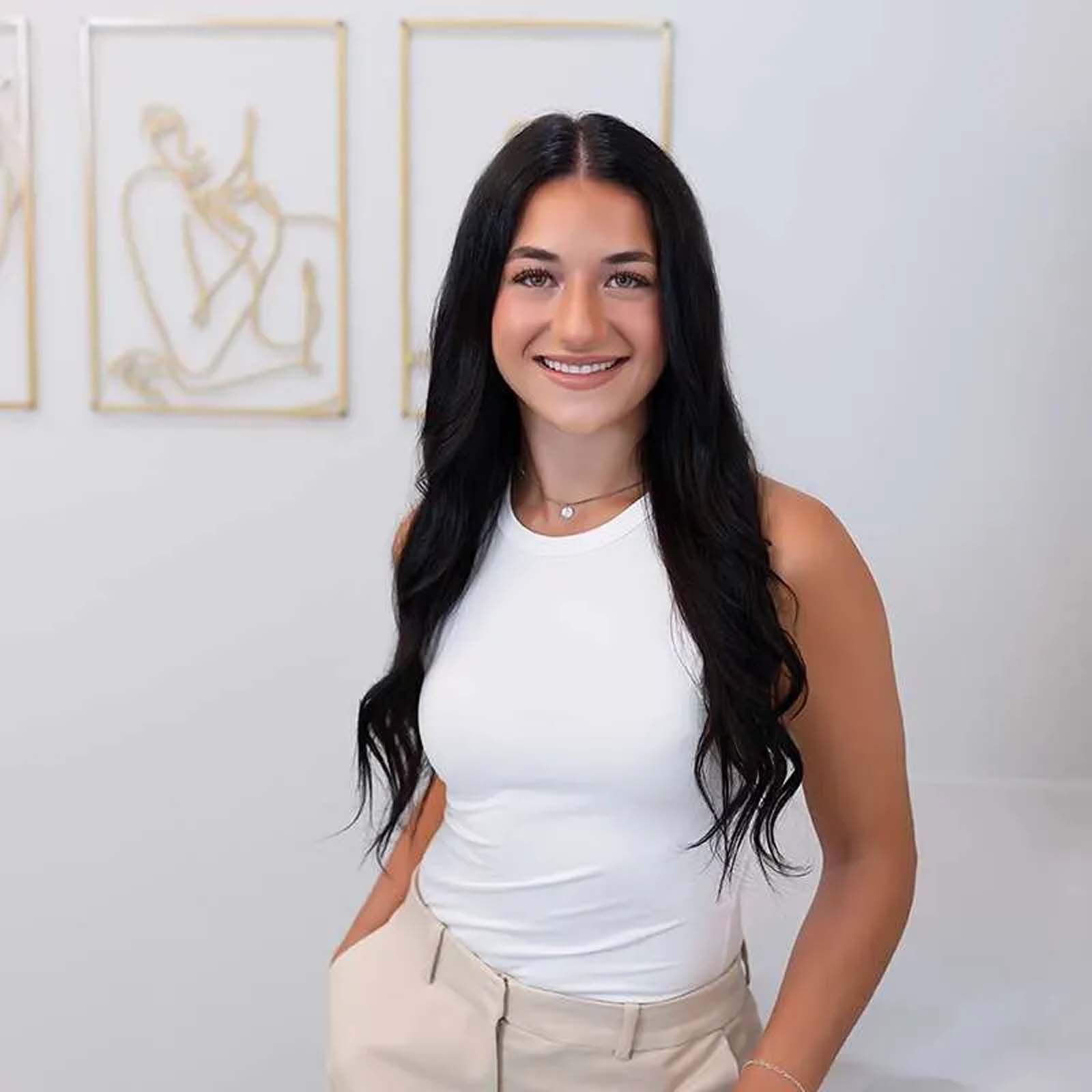 A woman with long dark hair is wearing a white tank top and khaki pants.