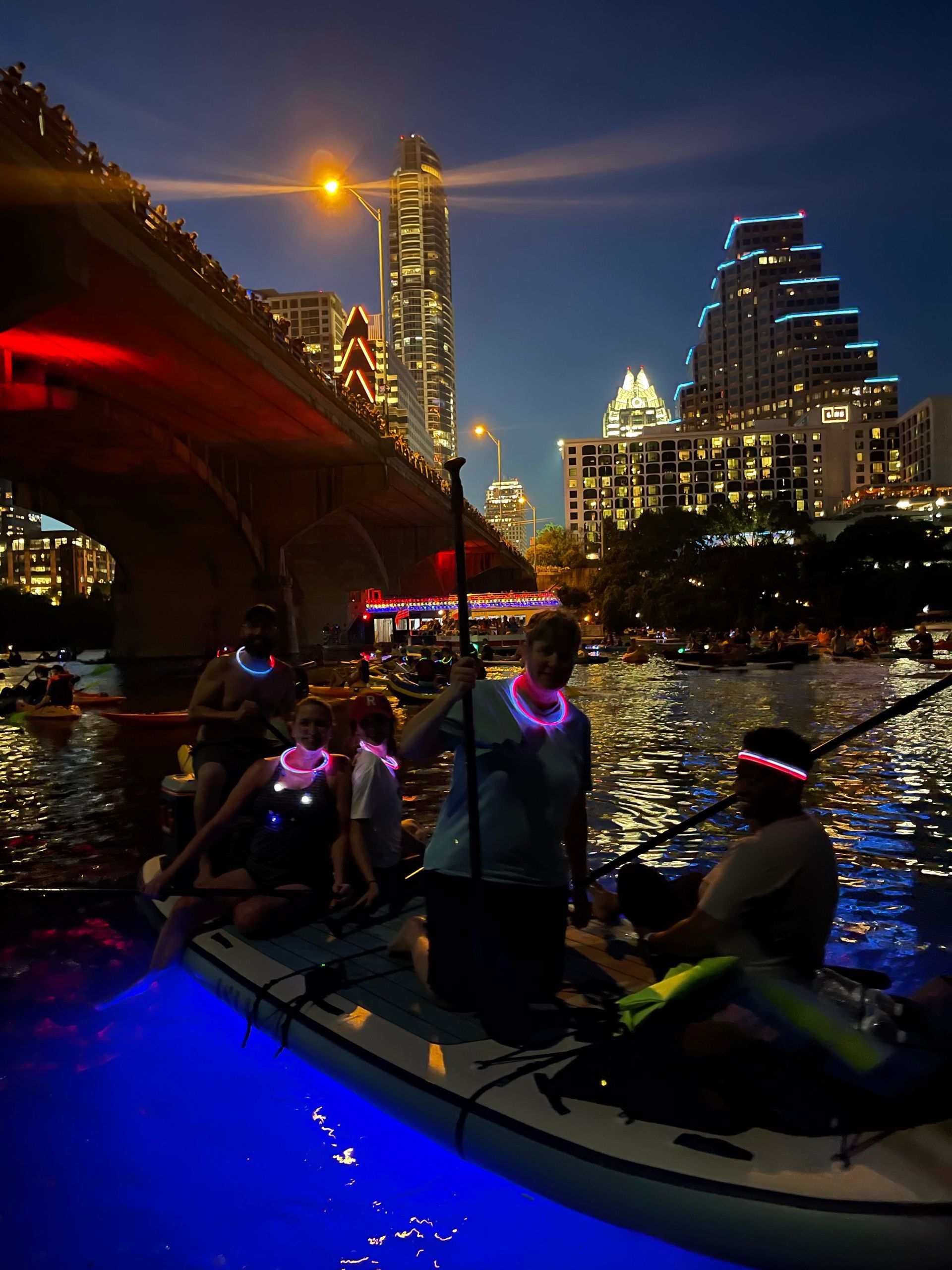 Group of friends paddleboarding at night, laughing and enjoying the glow tour with the city skyline in the background.