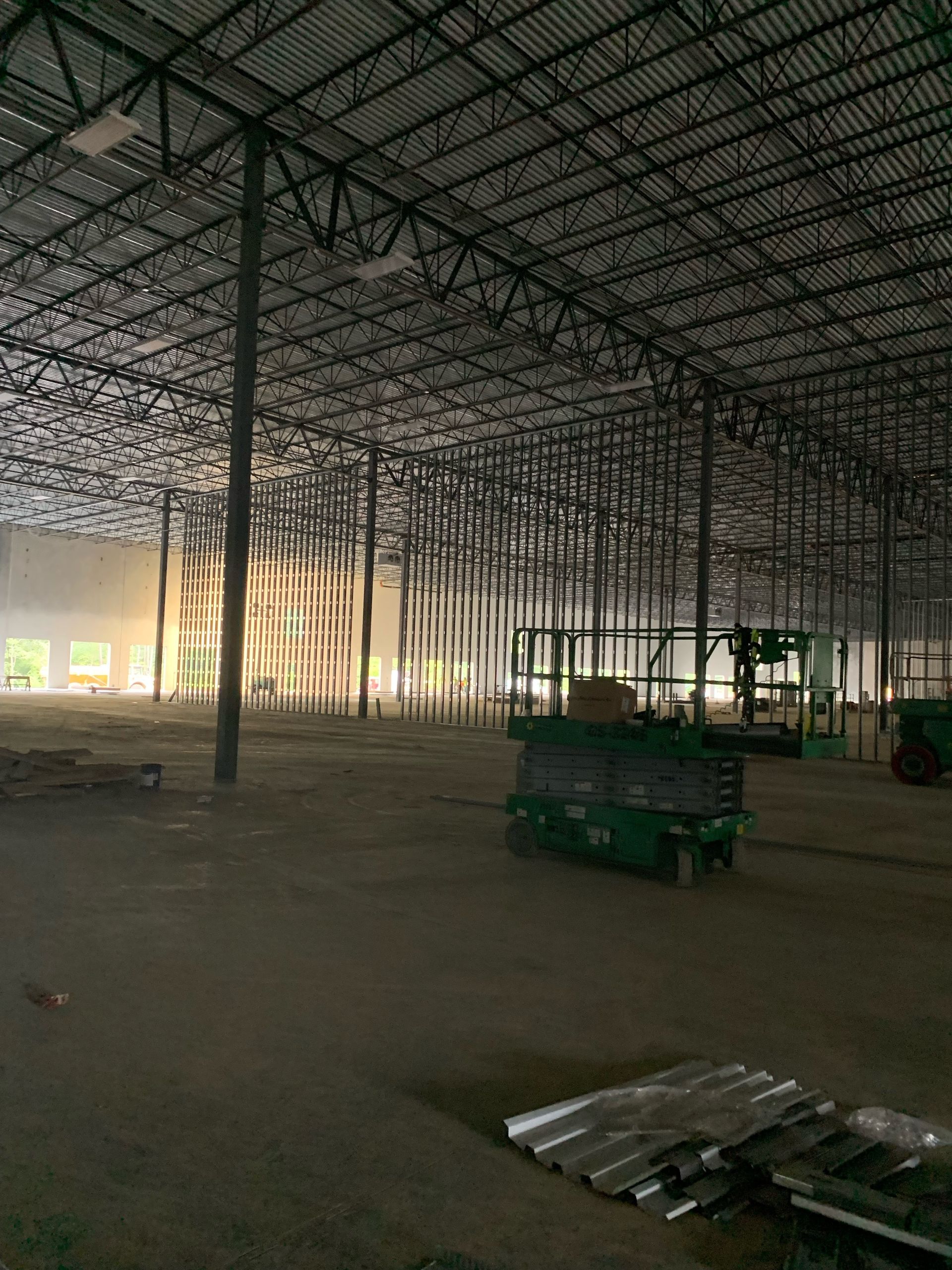 Large open warehouse interior with steel roof trusses, scattered carts, and stacks of metal sheets on the floor.