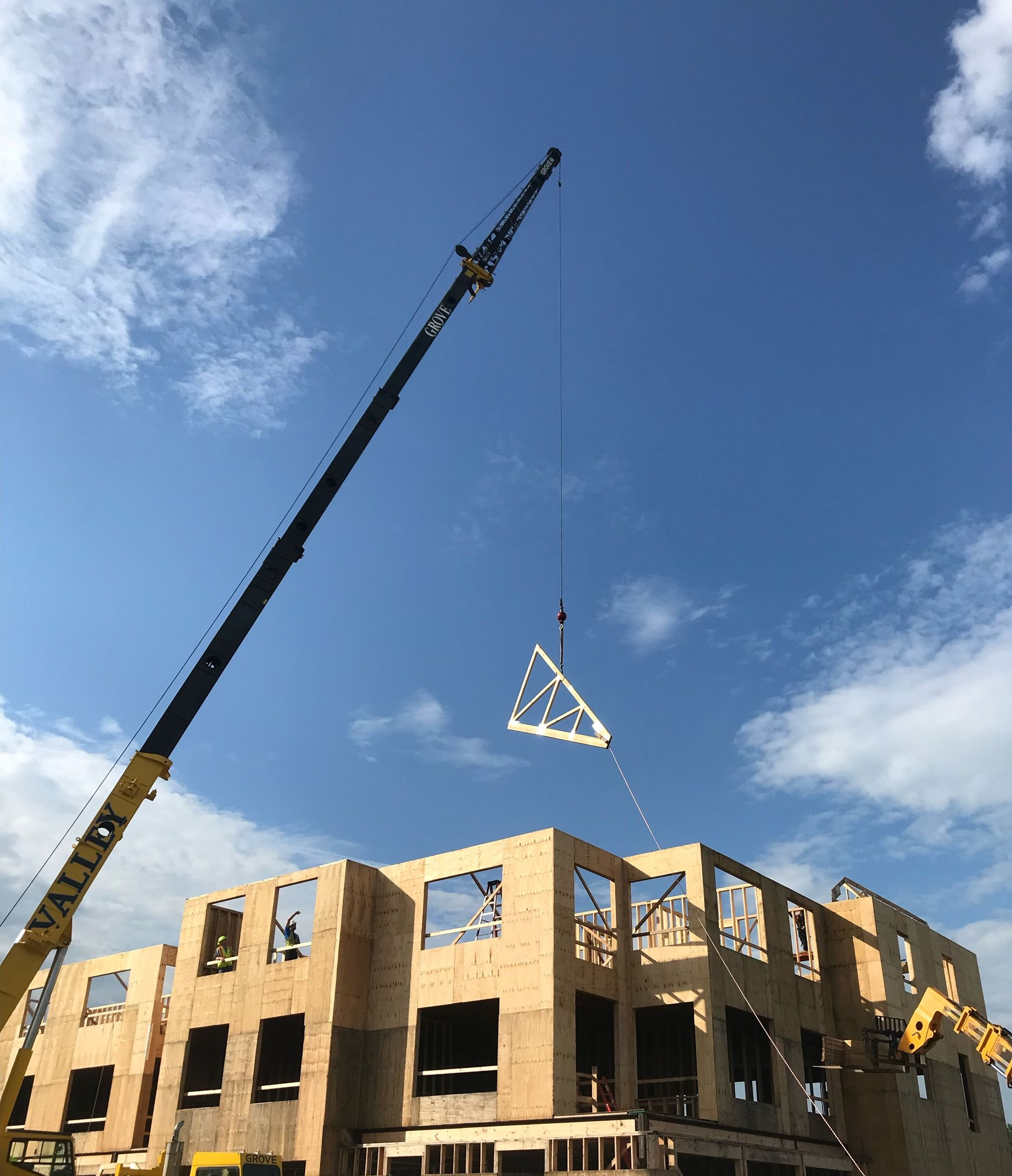 Construction site with brick building, crane lifting truss, and blue sky with clouds
