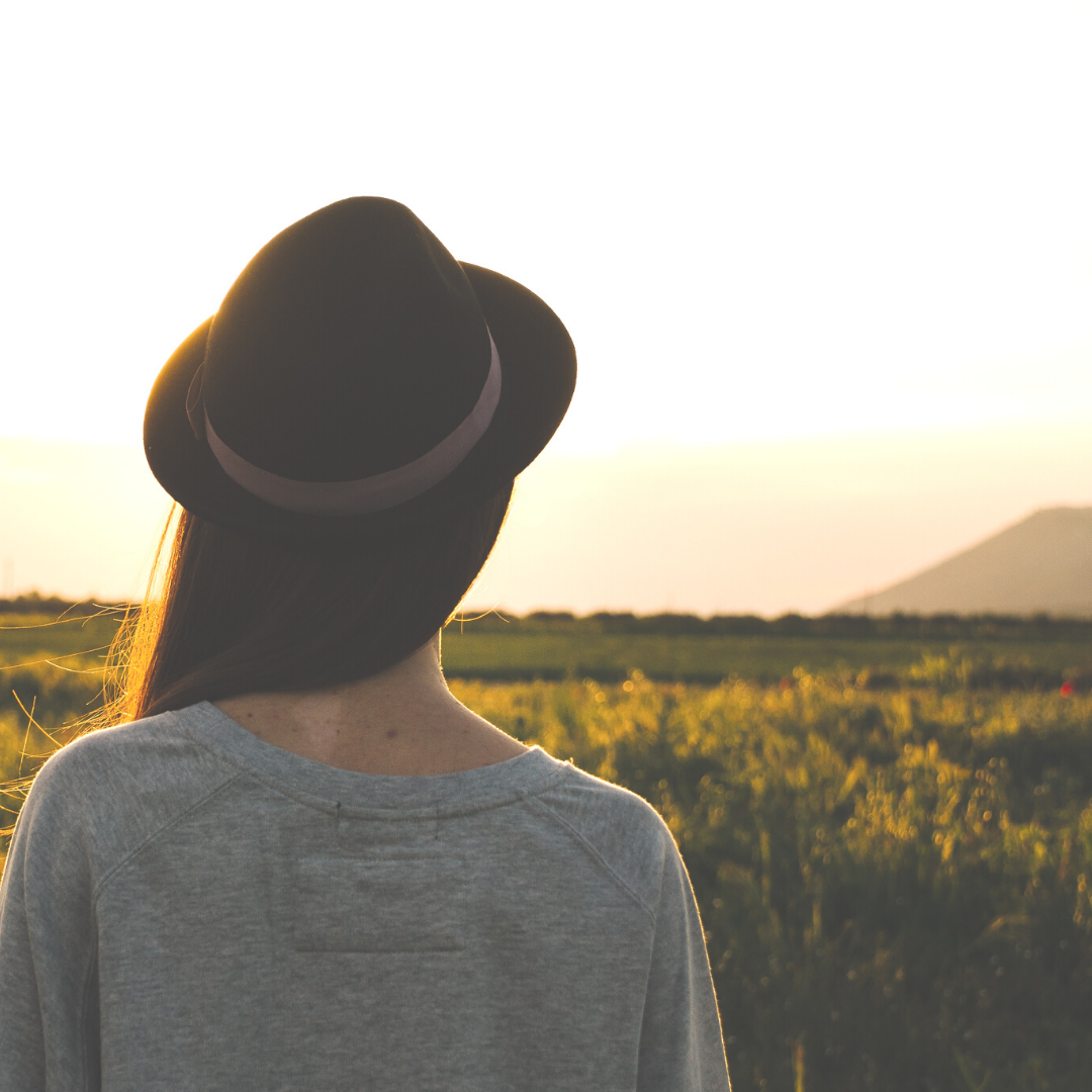 Viewed from the back, a woman with long brown hair and wearing a brown hat looks to the right at the horizon.