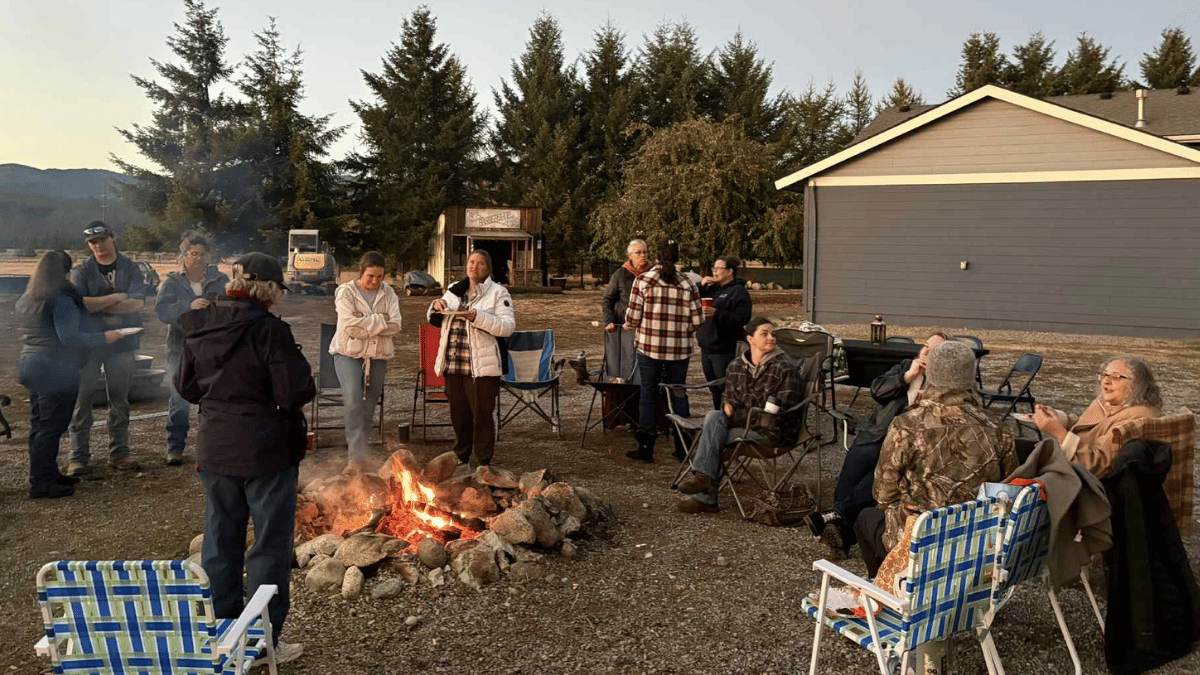 women gathered around a bonfire outdoors near a building and trees at dusk.