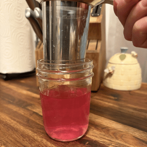 Hand straining bright pink vinegar into a glass jar on a wooden table