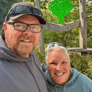 Smiling couple outdoors by the Polyface Farm wooden sign in a wooded area