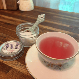 Pink tea in a floral cup and saucer beside a small jar and spoon on a wooden table.