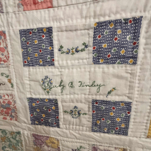 Close-up of a quilt block with blue patterned squares, floral embroidery, and the name “Ruth B. Finley” stitched in green