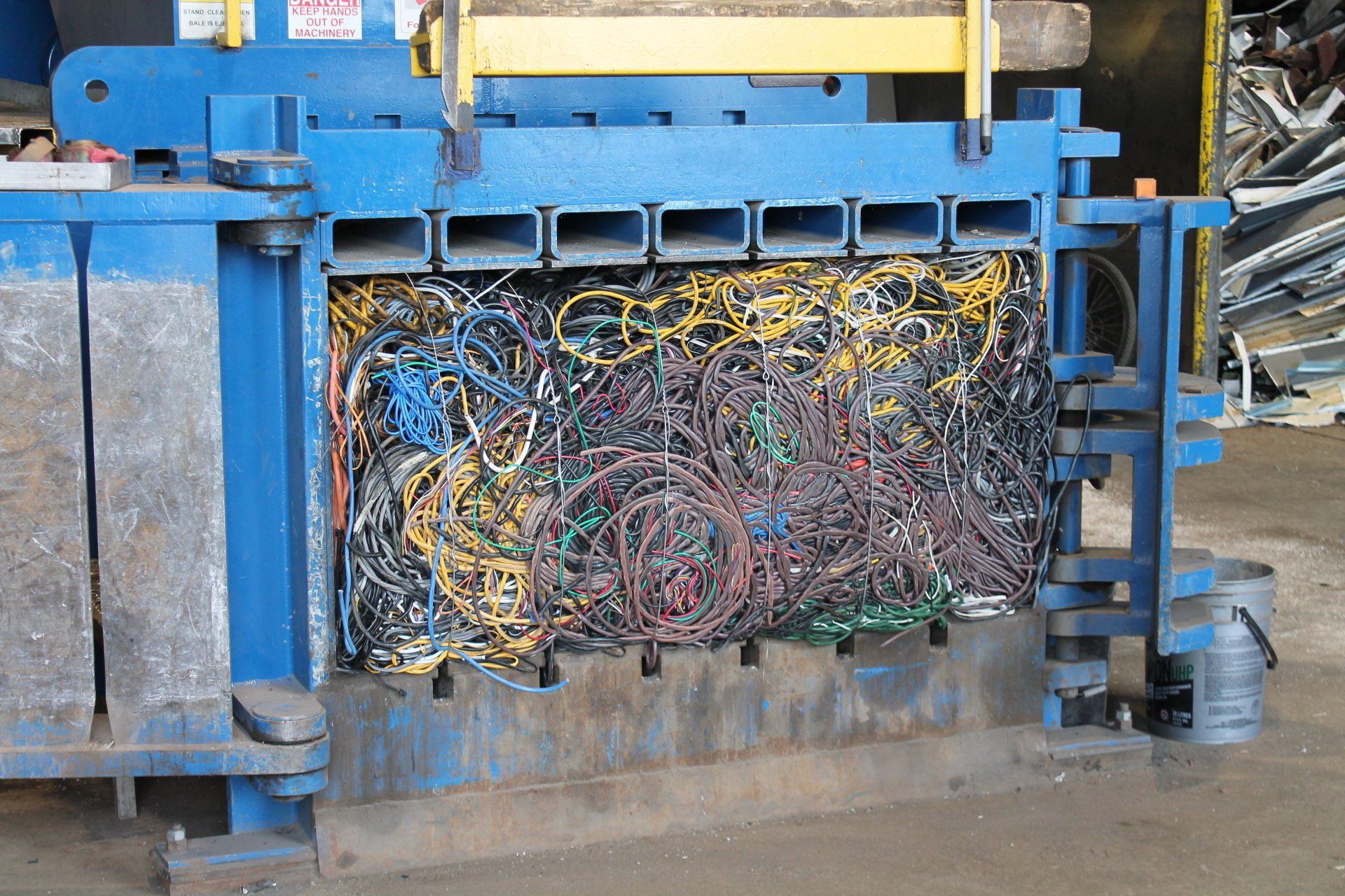 Blue industrial baler compressing a tangled bundle of scrap wires and cables in a factory setting