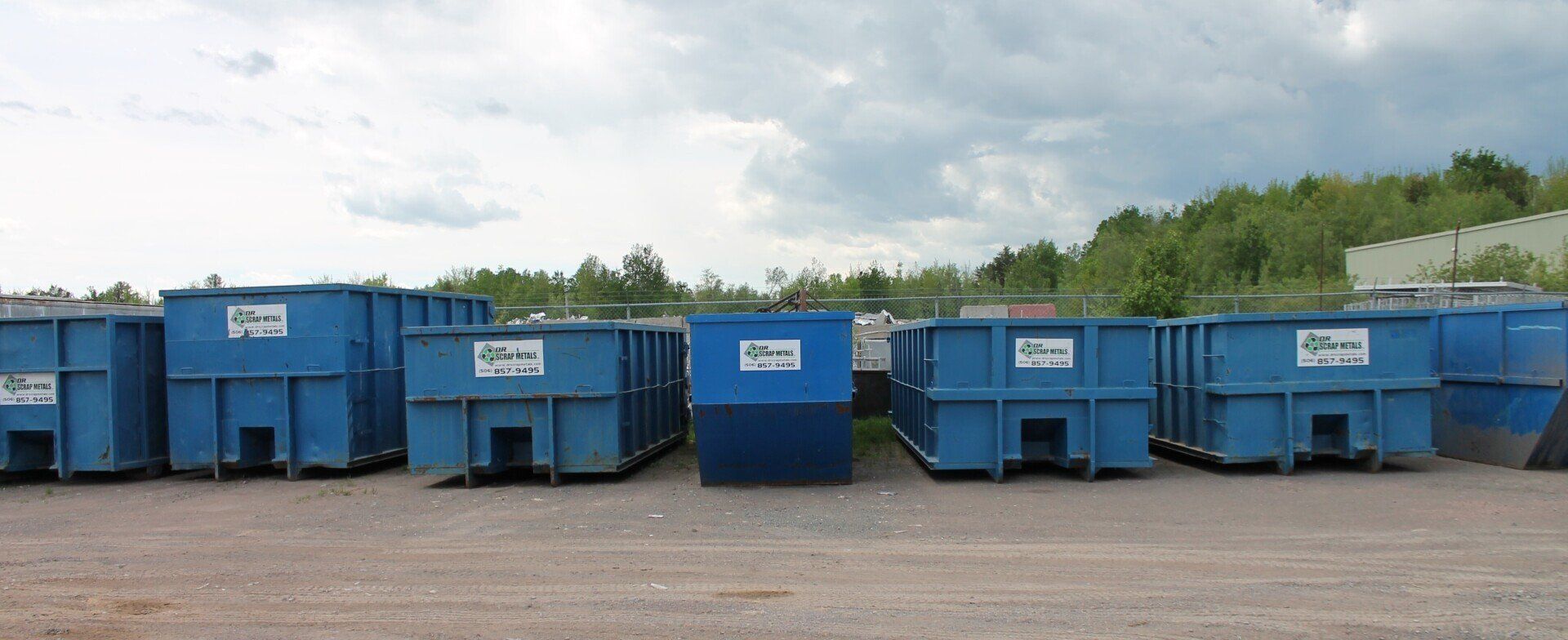 Blue industrial dumpsters lined up in a gravel lot under a cloudy sky