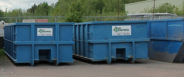 Blue industrial dumpsters in a lot, with greenery and a building in the background