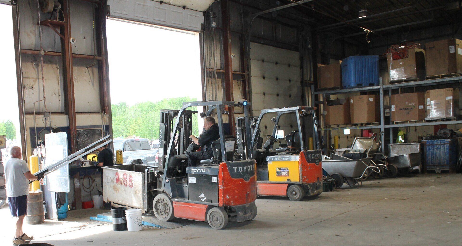 Forklifts in a warehouse with workers near a loading bay and shelving.