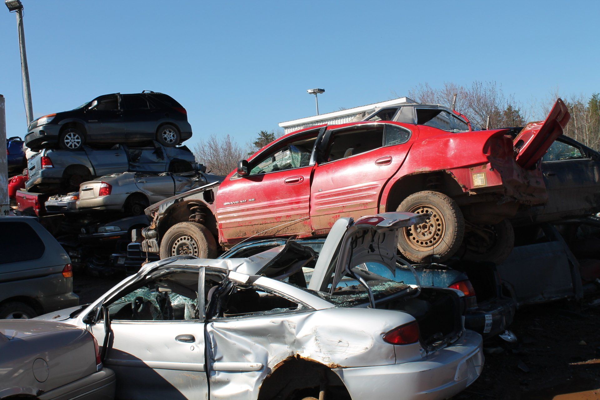 Stacked wrecked cars in an outdoor auto scrapyard, including a red SUV and a crushed white sedan.