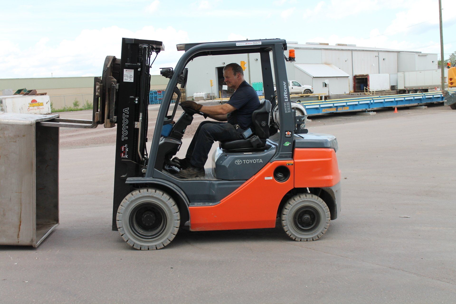 Orange forklift with a seated operator moving a large metal bin in an industrial yard