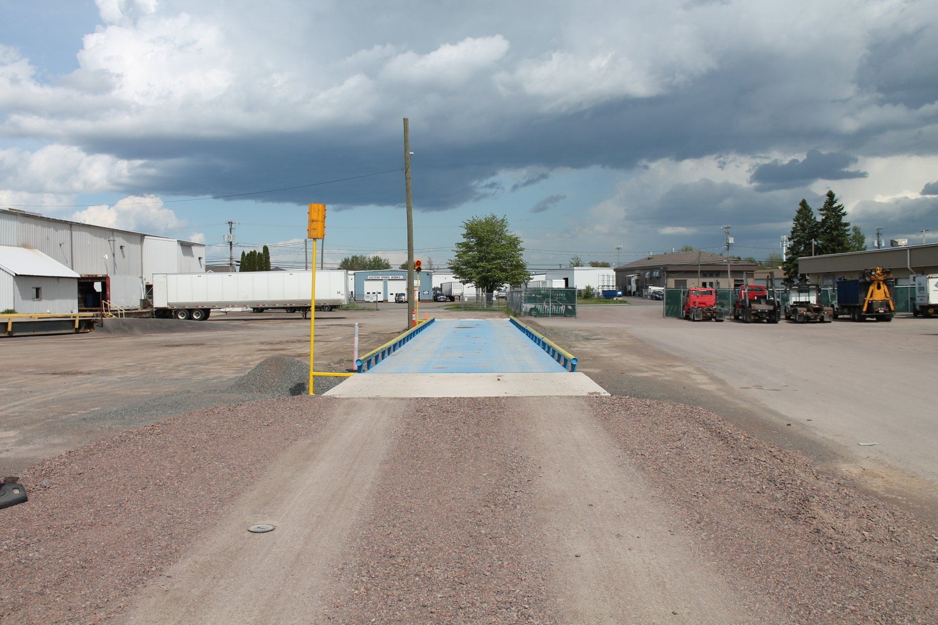 Blue raised walkway with yellow railings on a gravel lot under dark storm clouds, industrial buildings nearby