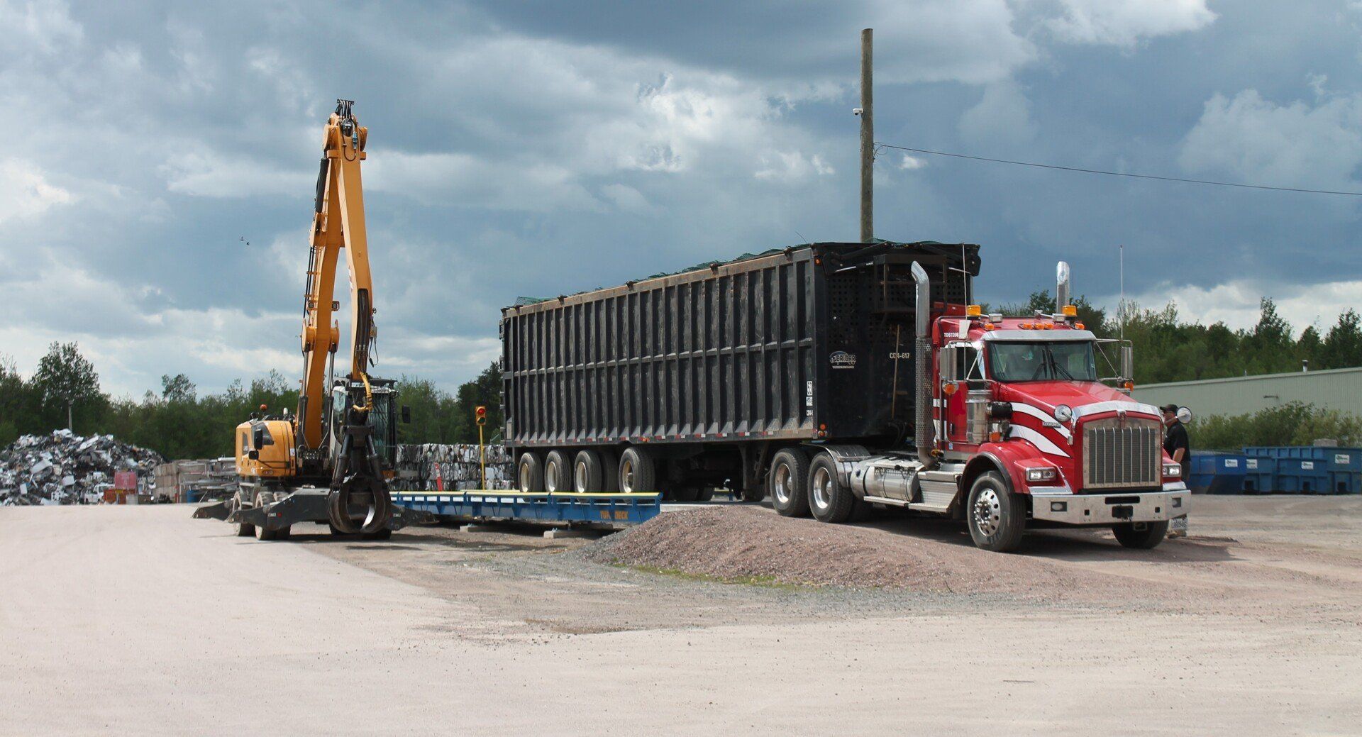 Red semi truck hauling a large black dumpster-like container beside an excavator in a gravel lot under cloudy skies
