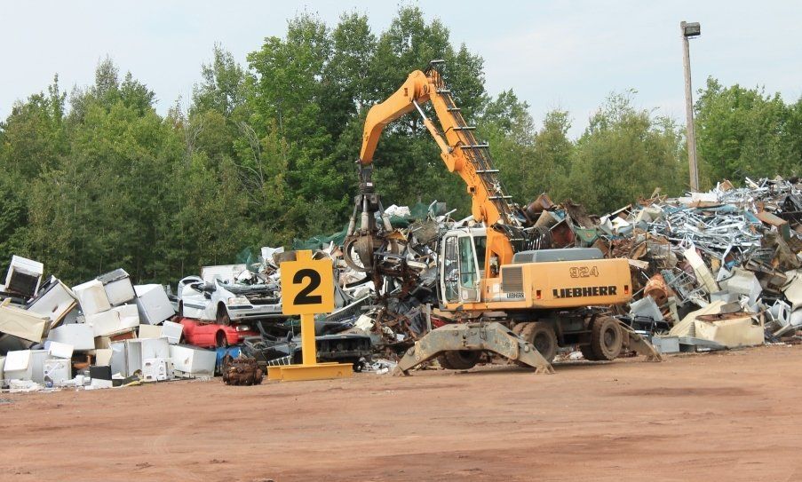 Yellow excavator sorting scrap metal in a junkyard, with piles of debris and trees in the background