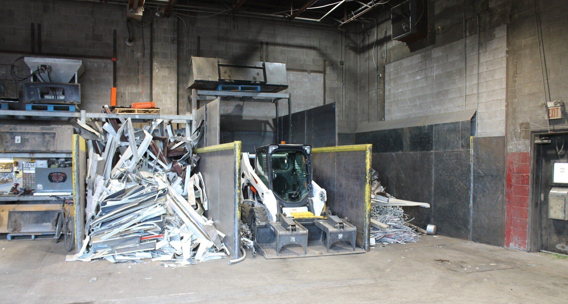 Industrial warehouse with a baler, piles of crushed metal scrap, and concrete walls.