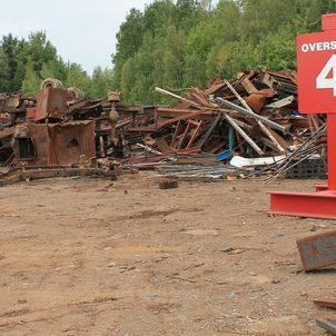 Rusty scrap metal piled in a dirt lot beside a red sign and trees.
