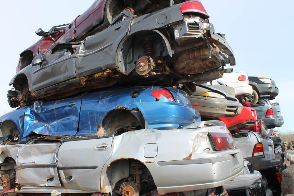 Stacked crushed cars in a scrapyard, including black, blue, and silver vehicles.