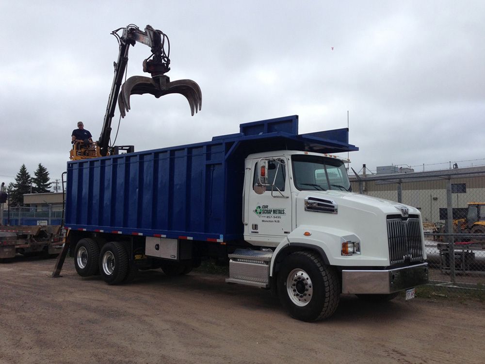 White truck with blue dump bed and crane lifting scrap in an industrial yard