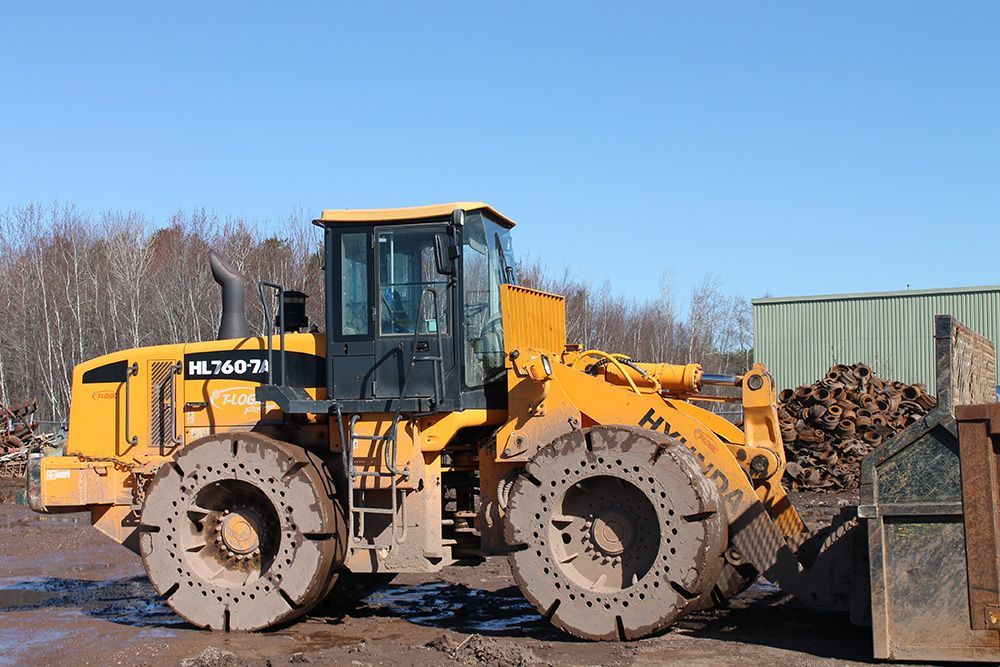 Yellow front-end loader on a muddy lot beside a woodpile and shipping container under a blue sky