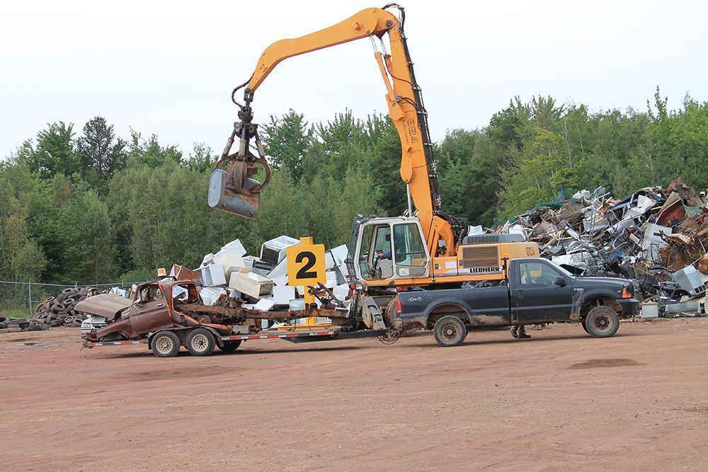 Excavator lifting scrap metal in a junkyard with piled wrecked cars and trucks nearby.