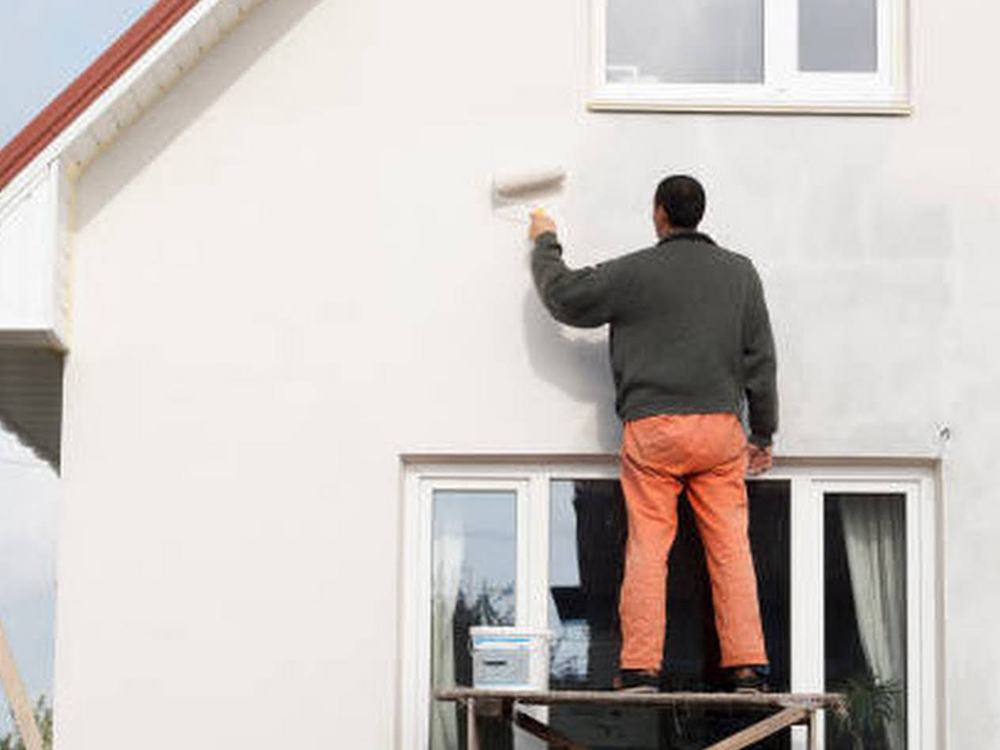 A man is standing on a scaffolding painting the side of a house.