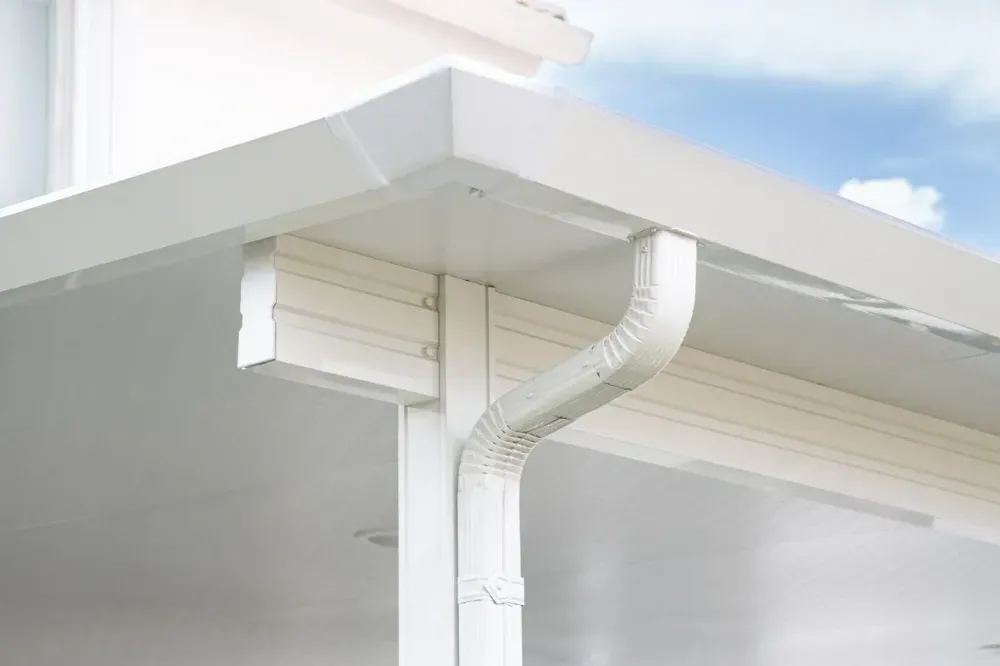 White rain gutter and downspout on a house roof corner against a blue sky