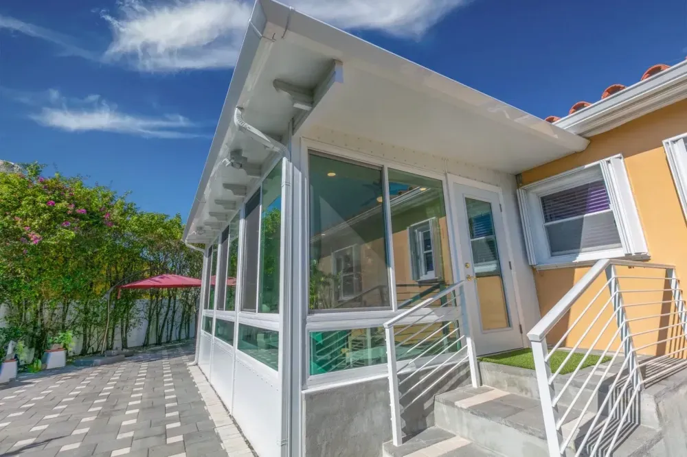 Sunlit patio with glass enclosure, white railing, and yellow house exterior under a blue sky