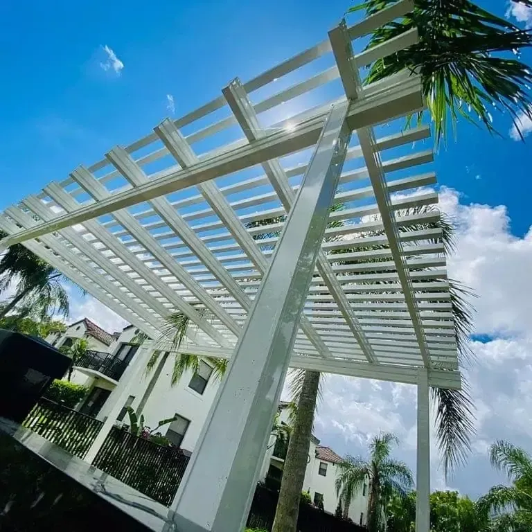 White pergola walkway beside modern white buildings, framed by palm trees under a bright blue sky