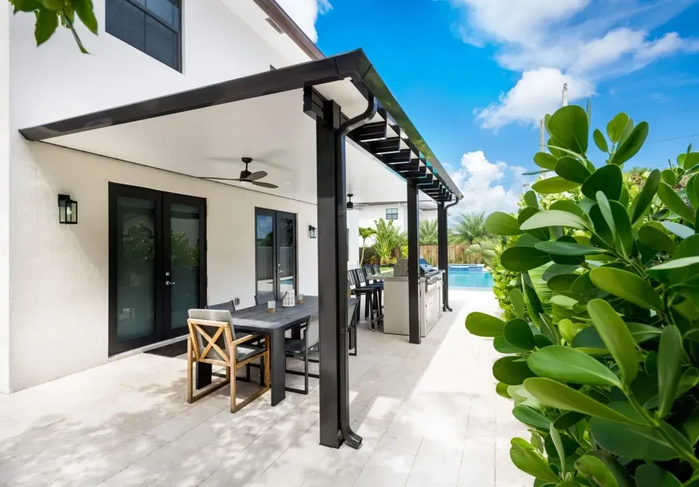 Modern patio with black dining table, sliding glass doors, white walls, and lush green plants under a blue sky