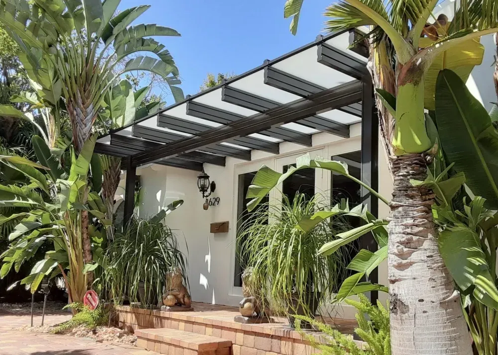 White house with a black pergola, surrounded by lush tropical plants and palm trees.